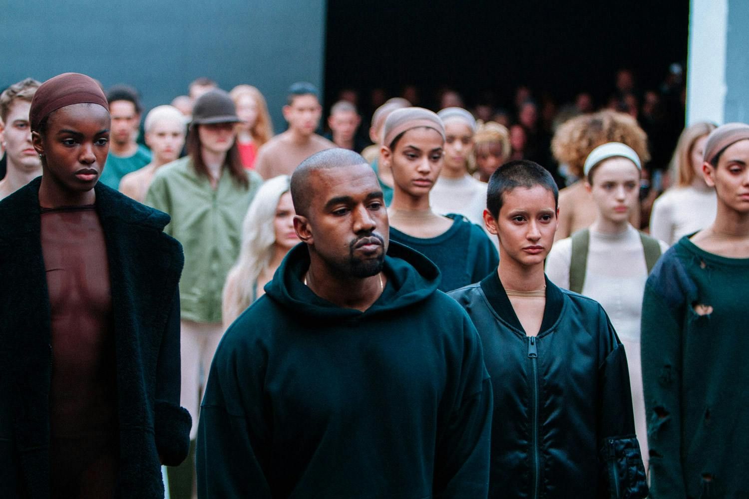 Yeezy Season 1 had Beyoncé front row. Ye bowed in a plain hoodie.. Ye in a dark hoodie standing among the models at the end of Yeezy Season 1, Skylight Clarkson Sq, New York, 12 Feb 2015.