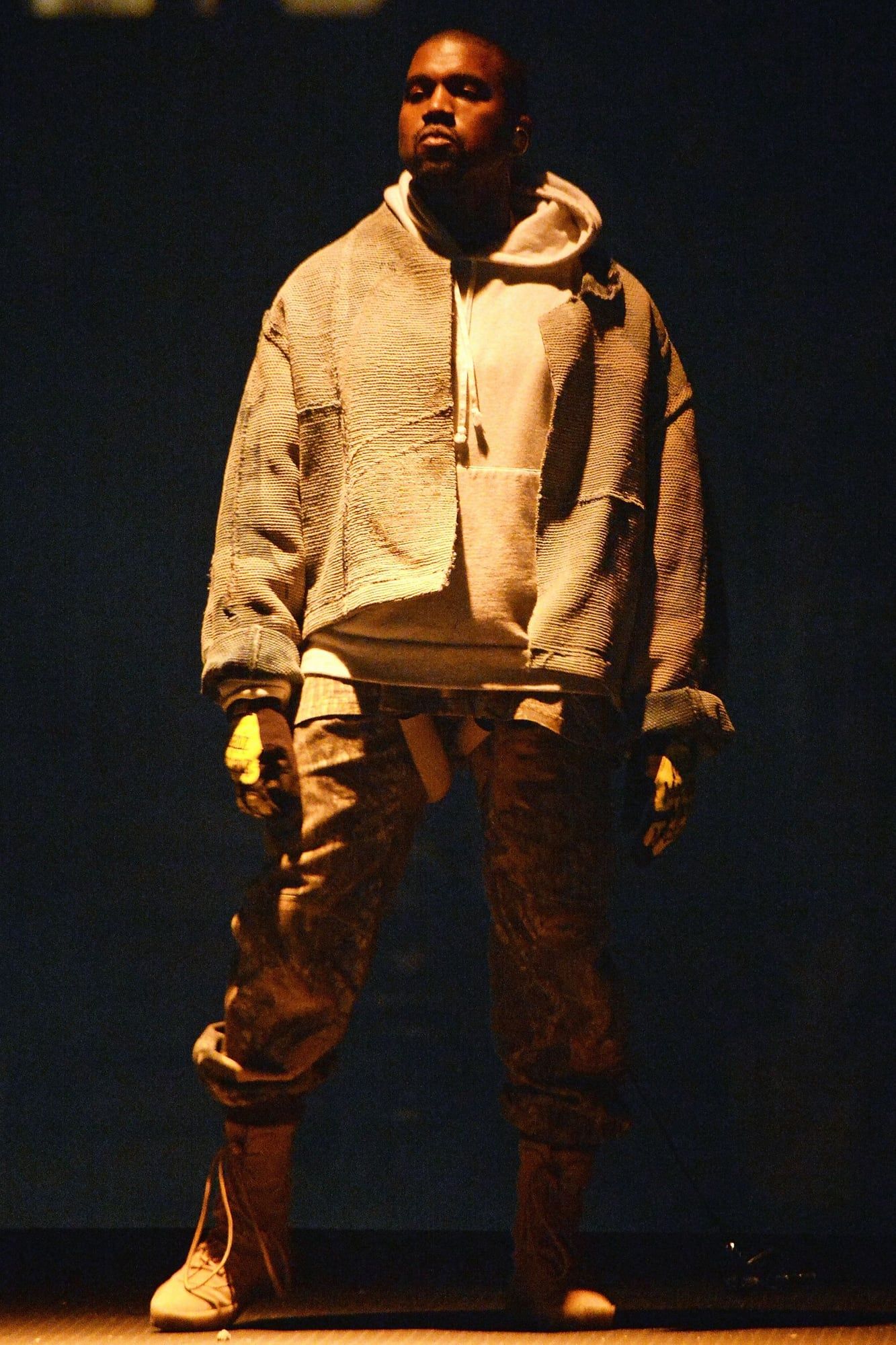 Ye’s Saint Pablo Tour had no stage, just a platform floating over the crowd. Ye full length on the Saint Pablo platform at Madison Square Garden, 5 Sep 2016: cropped textured jacket over a cream hoodie, camo trousers, tan lace-up boots, gloves.