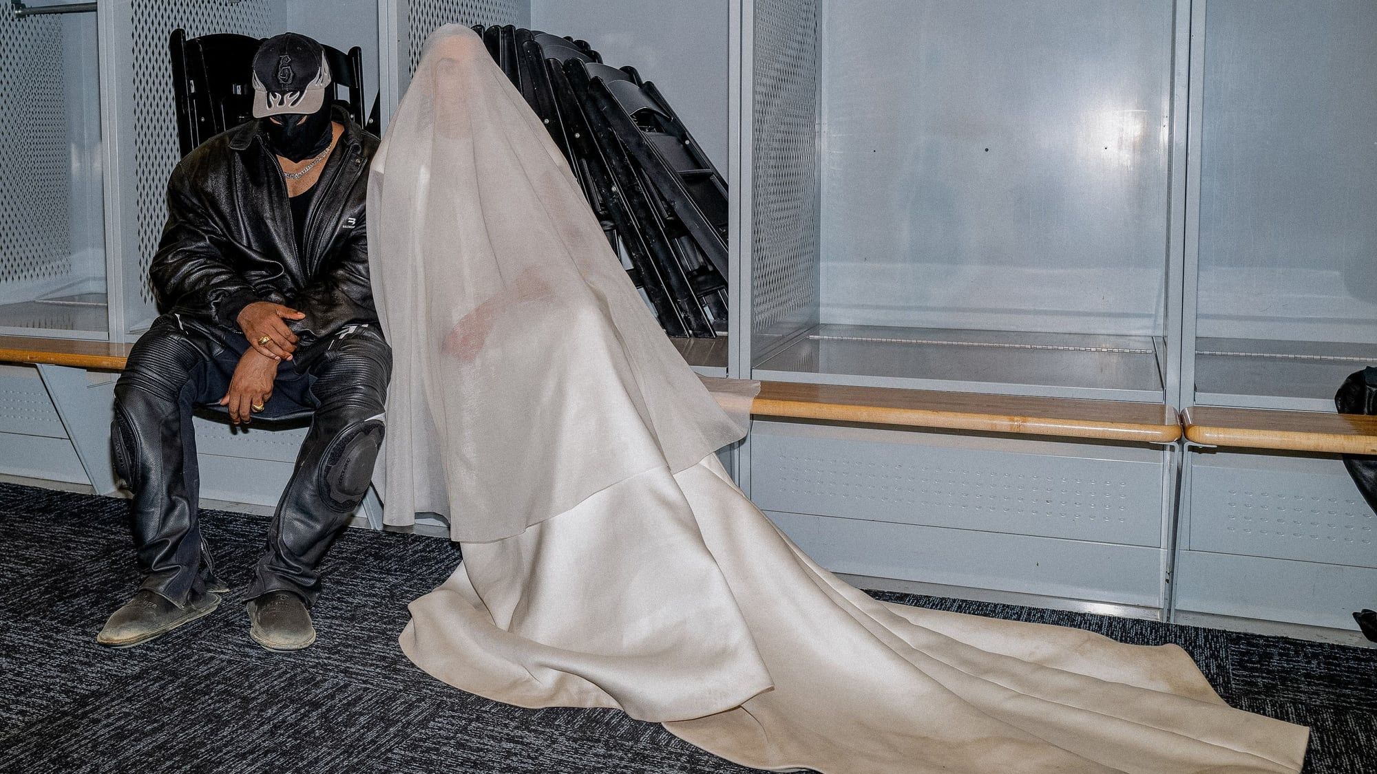 Ye rebuilt his childhood home inside Soldier Field, then walked out of it on fire. Ye backstage at Soldier Field in a flame-brim cap, black face covering, black Balenciaga leather jacket and leather biker trousers with Balenciaga knee pads, seated beside Kim Kardashian in the veiled Balenciaga couture gown, 26 Aug 2021.