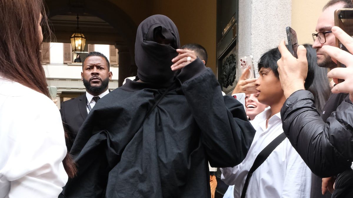 Ye walked Italy in socks all summer. By Christmas, you could order a pair. Ye in Milan on 22 Sep 2023, head wrapped in a black hood, oversized black top, fans filming on their phones.