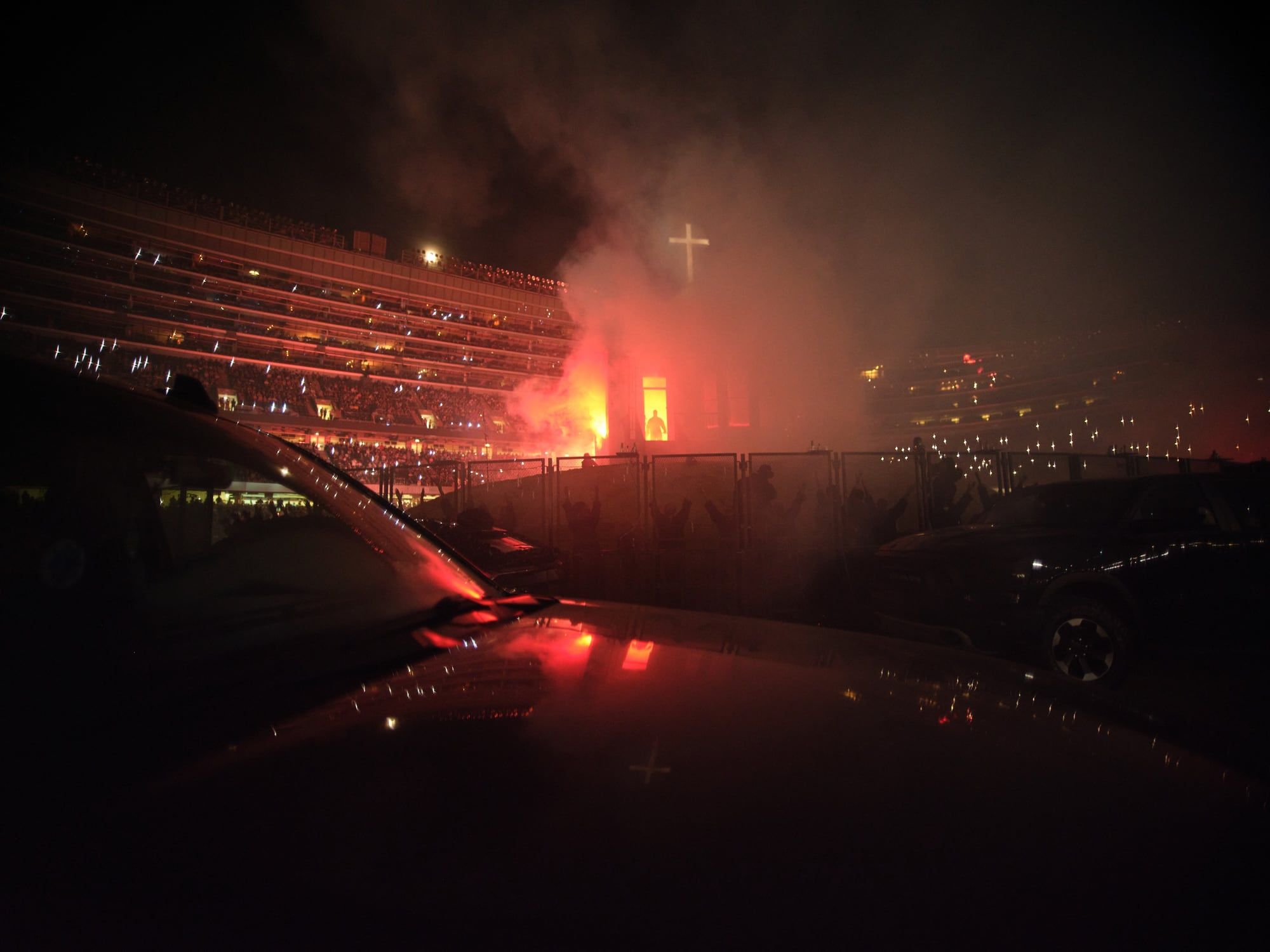 Ye rebuilt his childhood home inside Soldier Field, then walked out of it on fire. The replica house burning under the lit cross, a figure in the lit window, SUVs in the foreground, Soldier Field, 26 Aug 2021.
