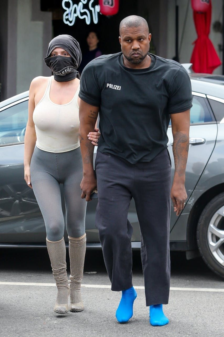 Ye walked Italy in socks all summer. By Christmas, you could order a pair. Ye in bright blue sock shoes, black Polizei tee and black trousers, Los Angeles, 25 May 2023, four months before Milan.